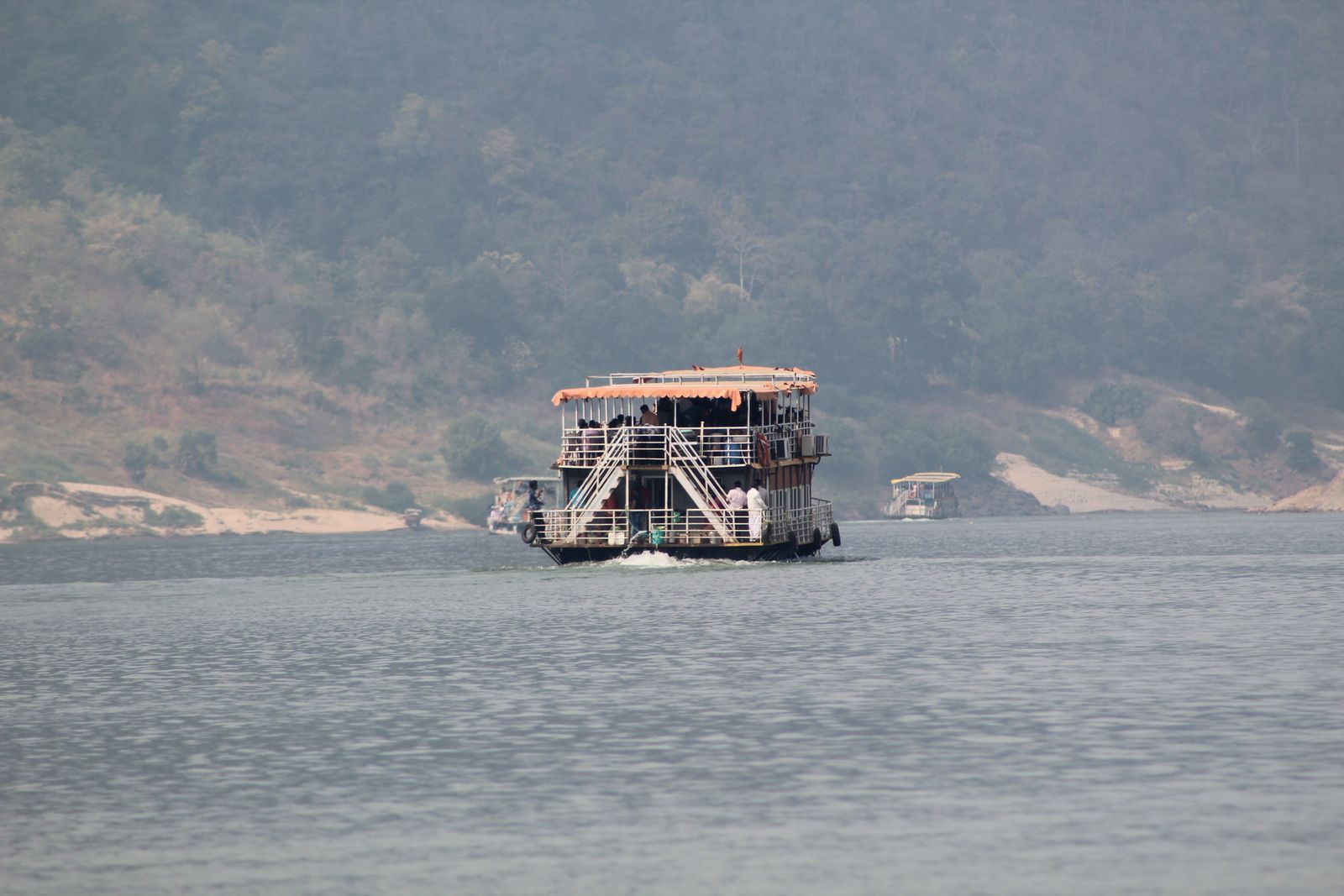 A double-deck passenger boat crossing a wide, hazy river beneath forested hills, similar to the craft used on Guwahati river trips