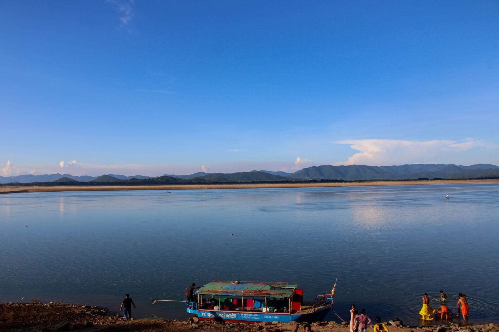 A painted wooden passenger boat at the edge of a broad, calm river in India with blue hills on the far shore